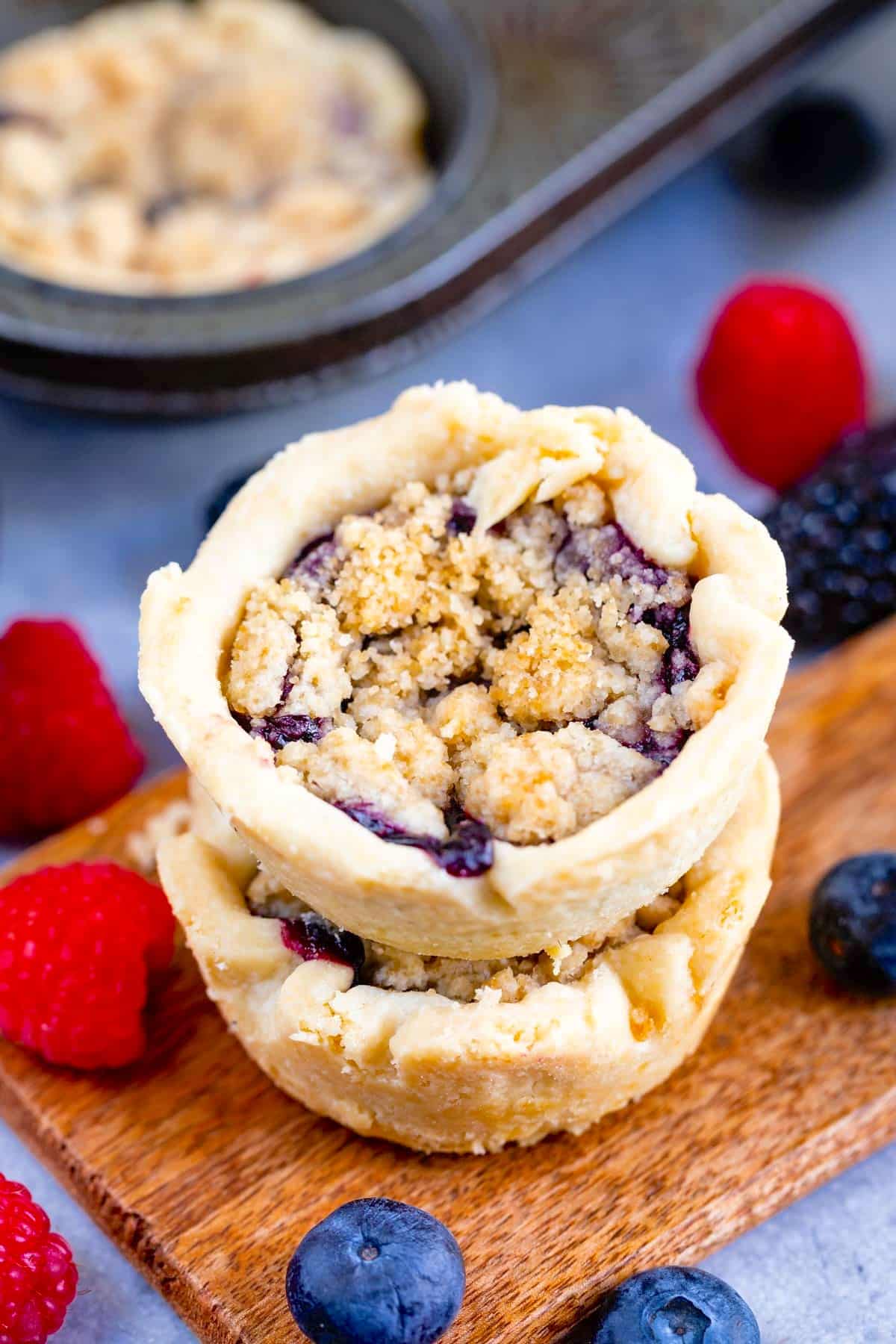 Deux mini tartes empilées sur une planche de bois, entourées de framboises fraîches, de myrtilles et de mûres, avec une plaque à pâtisserie et d'autres tartes en arrière-plan.