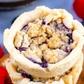 Close-up of a mini berry crumble pie with a golden, crumbly topping, displayed on a wooden surface. Fresh raspberries and blackberries are scattered in the background, highlighting the charm of homemade mini pies.