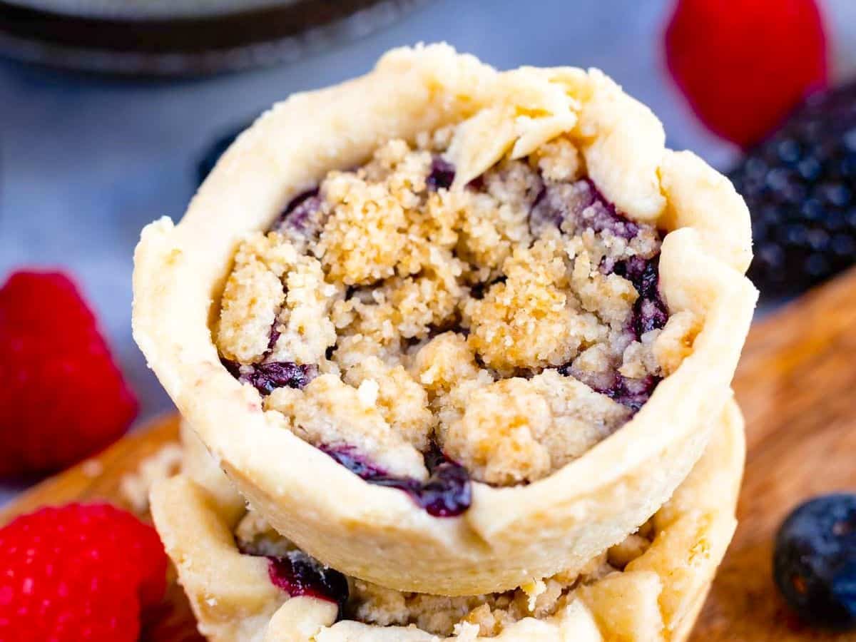 Gros plan d'une mini tarte crumble aux myrtilles avec garniture dorée, présentée sur une surface en bois. Des framboises et des mûres fraîches sont dispersées en arrière-plan, soulignant le charme des mini tartelettes maison.