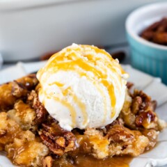 Pecan pie cobbler on a white scalloped dish topped with vanilla ice cream and caramel sauce