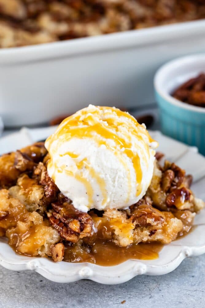 Pecan pie cobbler on a white scalloped dish topped with vanilla ice cream and caramel sauce