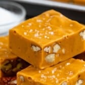 Close-up of rich butterscotch fudge squares with chopped pecans inside, topped with coarse sea salt. A small glass bowl of sea salt is visible in the background.