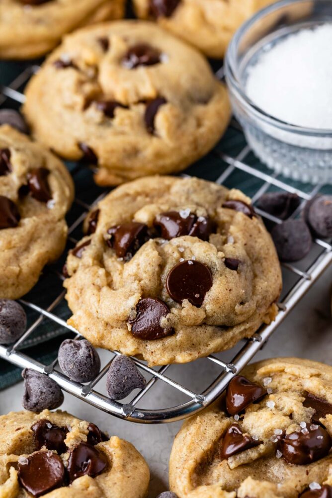 Salted chocolate chip cookies on a metal cooling rack with chocolate chips and a small dish of salt