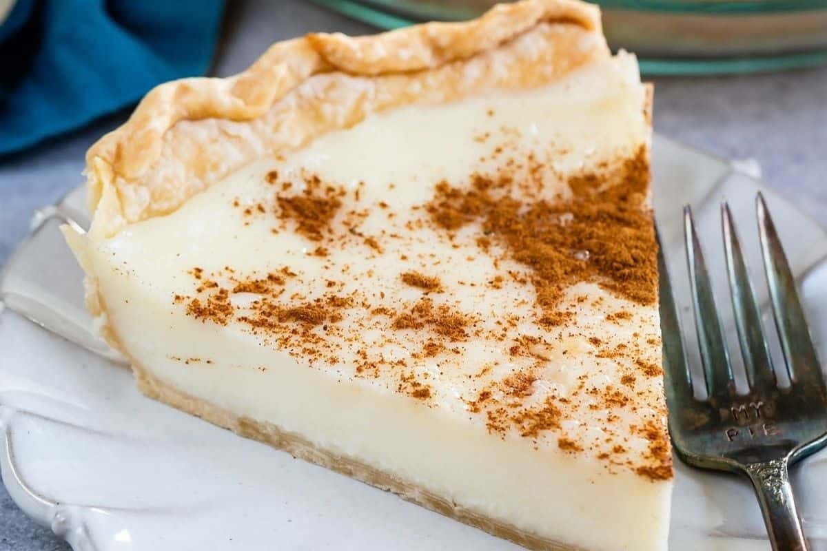 A slice of sugar cream pie dusted with cinnamon on a plate next to a fork.
