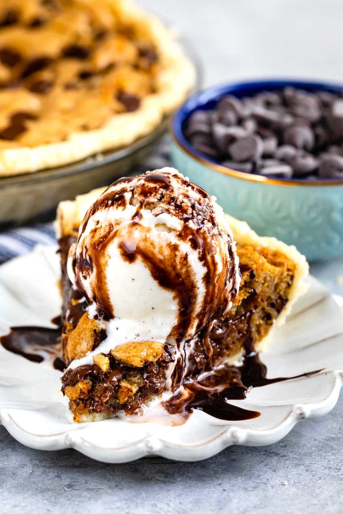 A decadent slice of chocolate chip cookie pie is topped with a scoop of vanilla ice cream and drizzled with rich chocolate sauce, served on a white plate with a bowl of chocolate chips in the background.