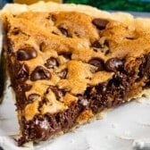 A close-up of a chocolate chip cookie pie slice showcases its golden crust, gooey chocolate filling, and generous chocolate chips on top, served on a white plate with a fork beside it.