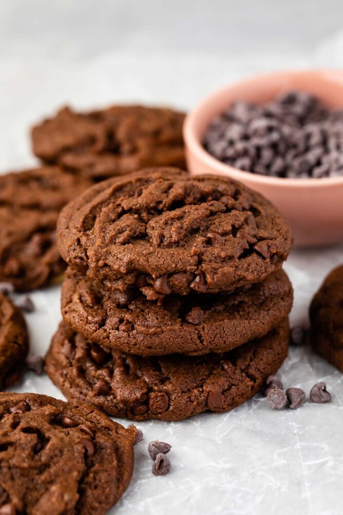 Three double chocolate chip cookies stacked on top of eachother with bowl of chocolate chips behind it