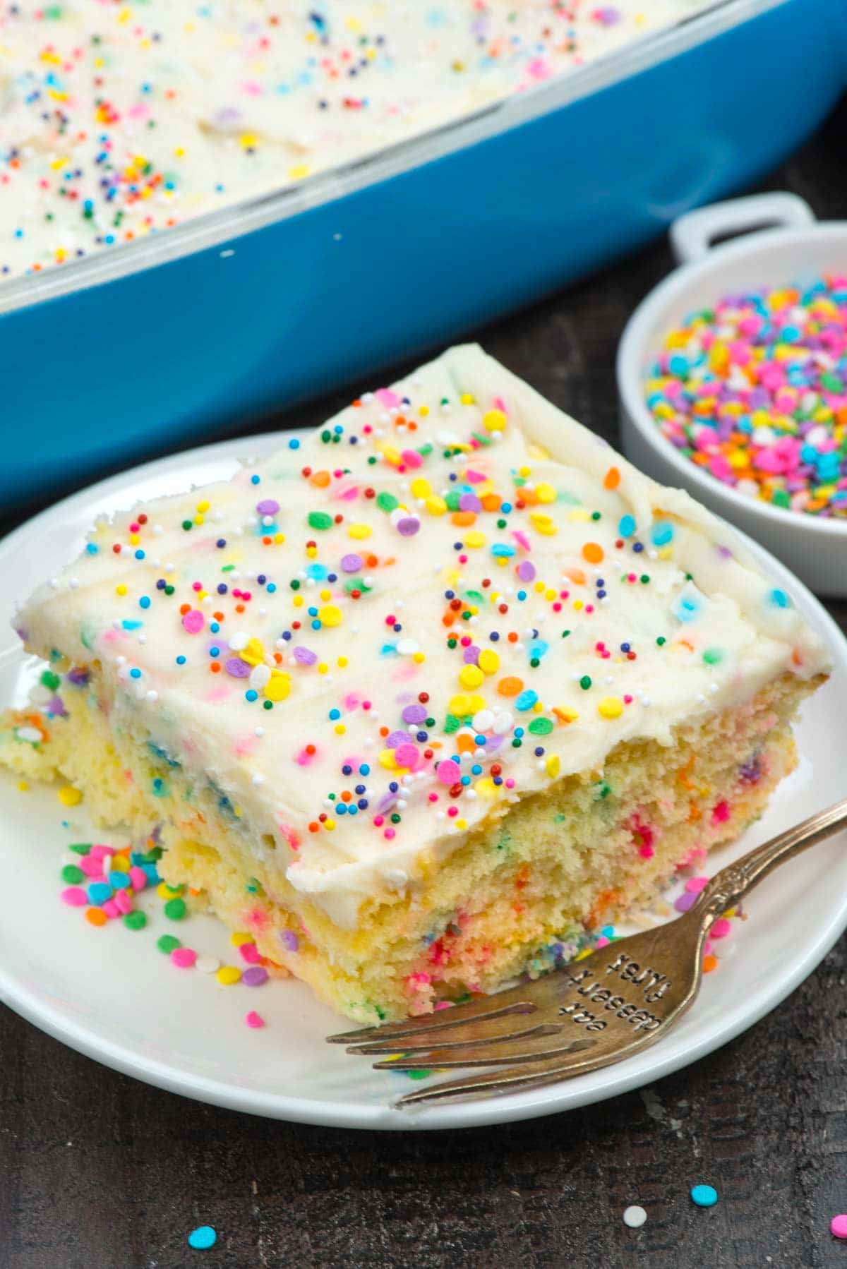 One square slice of homemade funfetti cake on plate surrounded by rainbow sprinkles