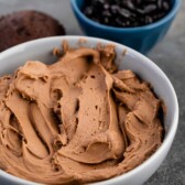 Mocha frosting in a bowl with chocolate cupcakes around it
