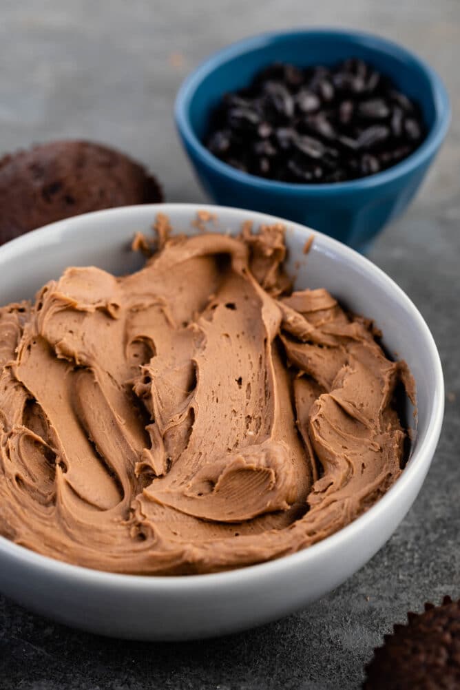 Mocha frosting in a bowl with chocolate cupcakes around it