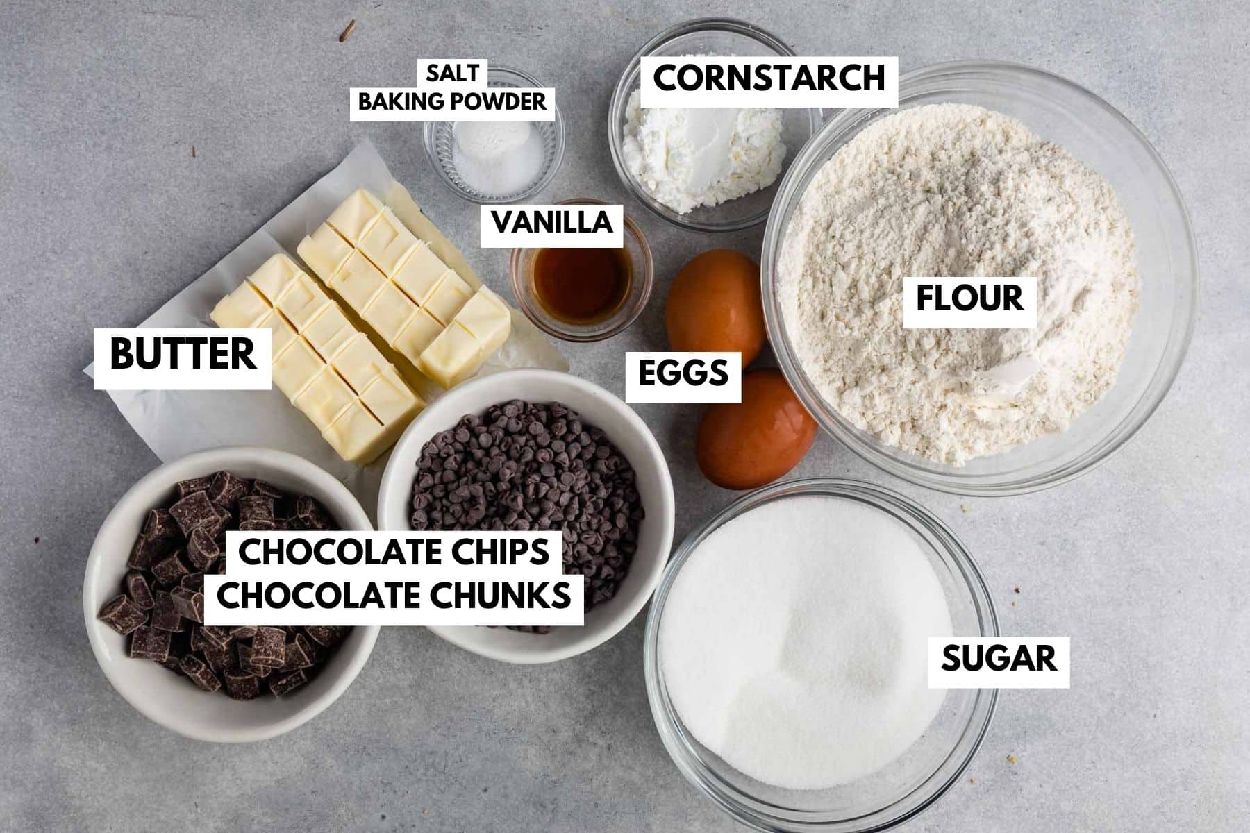 Overhead view of baking ingredients in bowls: butter, chocolate chips and chunks, sugar, eggs, flour, cornstarch, vanilla, and a small bowl of salt and baking powder—all labeled with text—perfect for whipping up your favorite Disney cookies at home.