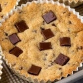 A close-up of a baked Disney cookie in a cupcake liner, topped with several square chunks of chocolate, on a textured baking sheet.