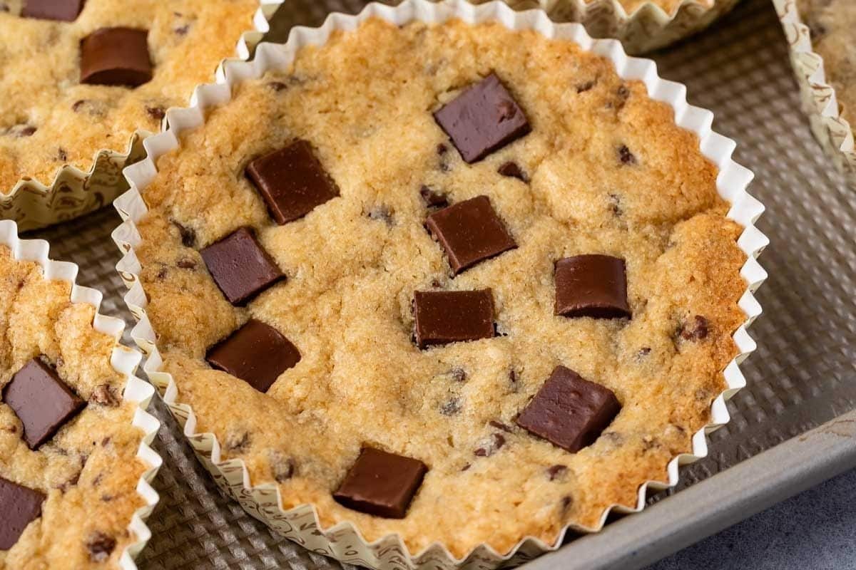 A close-up of a baked Disney cookie in a cupcake liner, topped with several square chunks of chocolate, on a textured baking sheet.