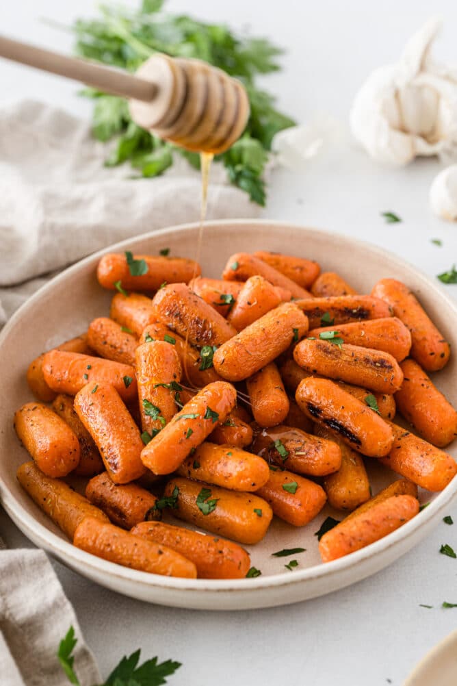 Roasted carrots in a white serving bowl with honey being poured over them