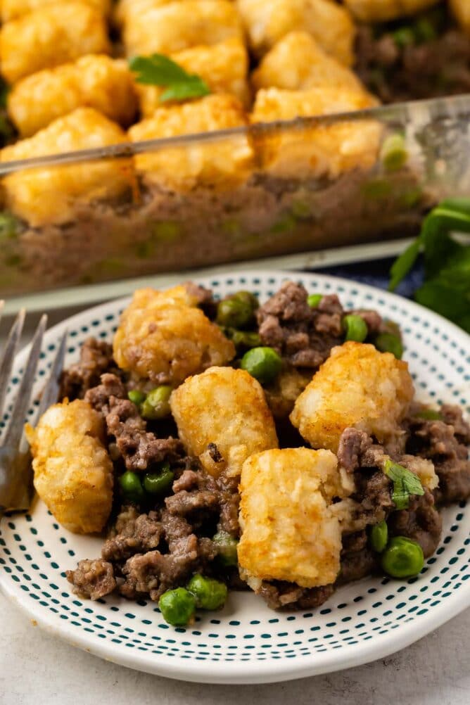 Hamburger tater tot casserole with gravy on a plate with full casserole dish in background