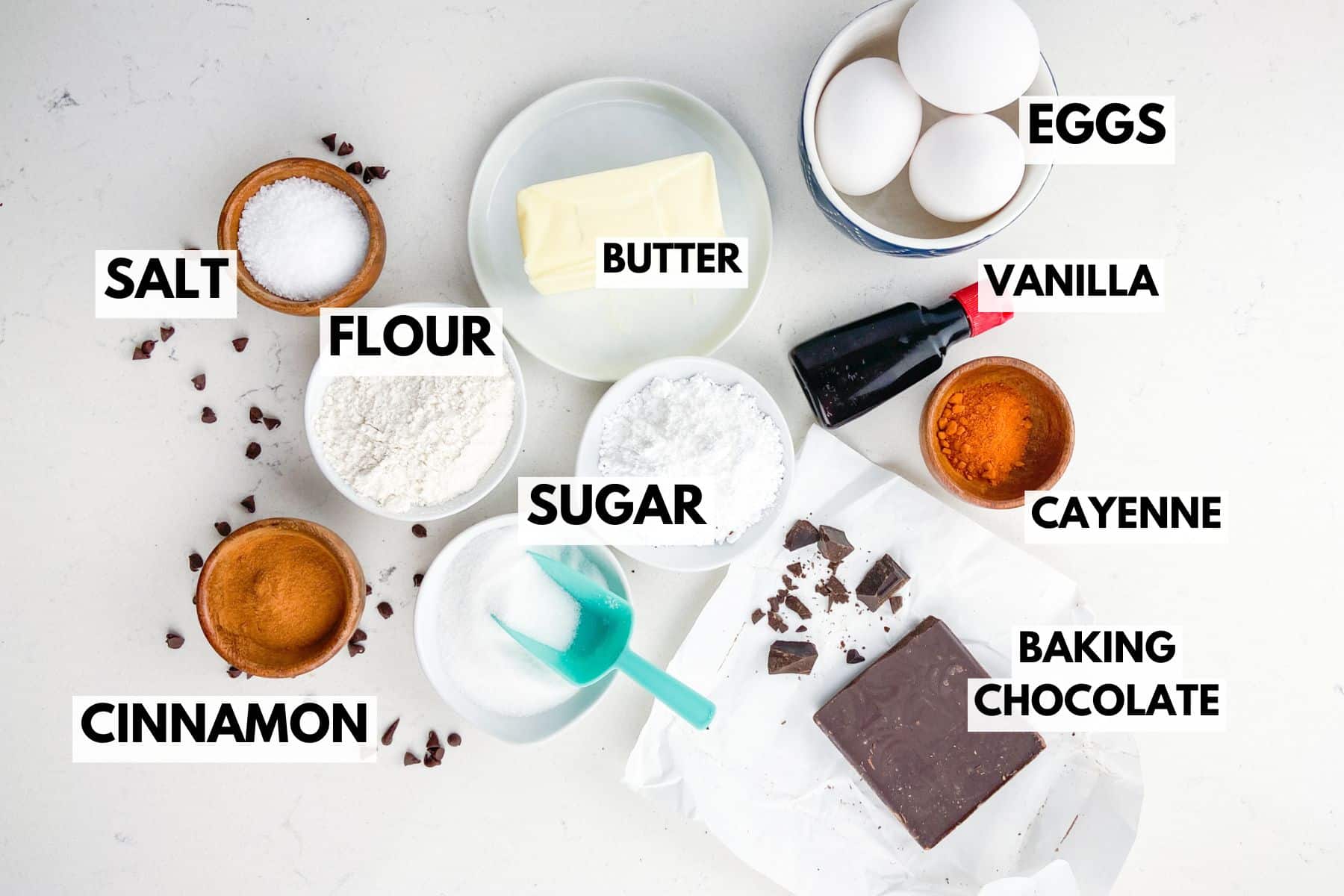 Overhead view of baking ingredients for Mexican Brownies on a white surface, each labeled: salt, flour, sugar, cinnamon, butter, eggs, vanilla, cayenne, and baking chocolate. Some chocolate pieces are unwrapped on paper.