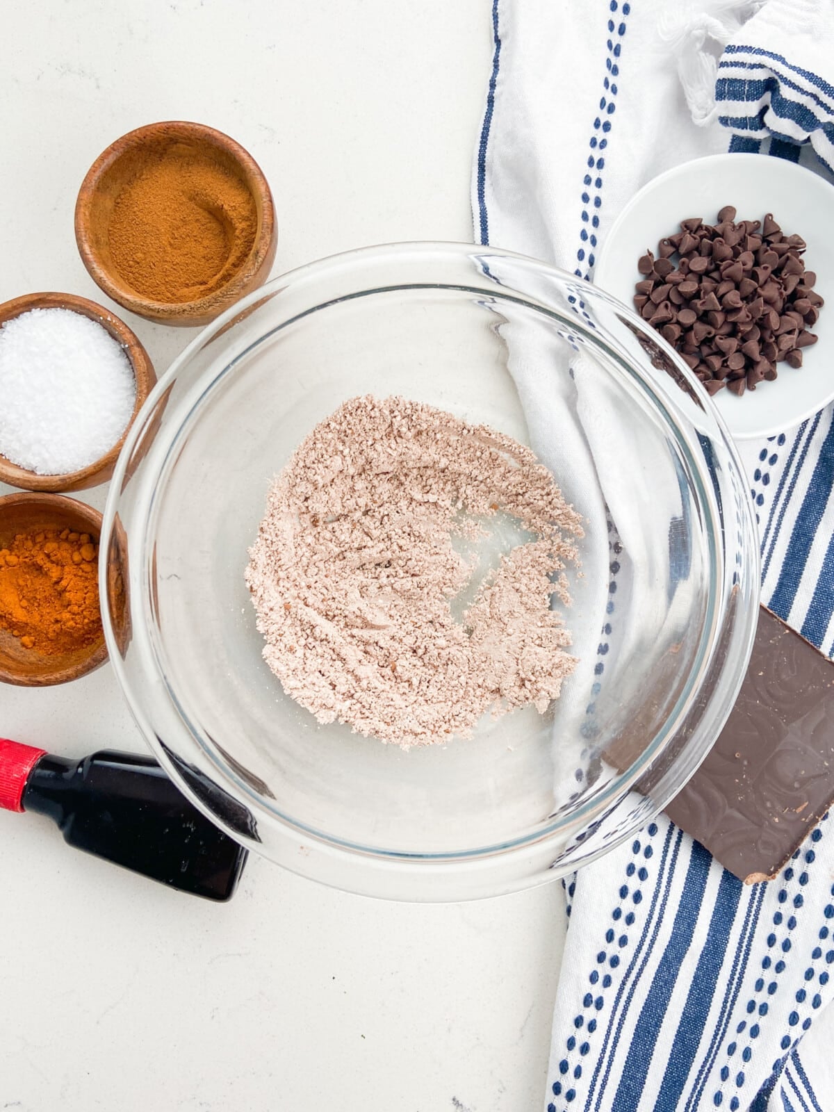 A glass mixing bowl with dry ingredients sits on a white counter, surrounded by small bowls of cinnamon, salt, chocolate chips, and a dark chocolate bar&mdash;perfect for whipping up flavorful Mexican Brownies&mdash;next to a striped kitchen towel.