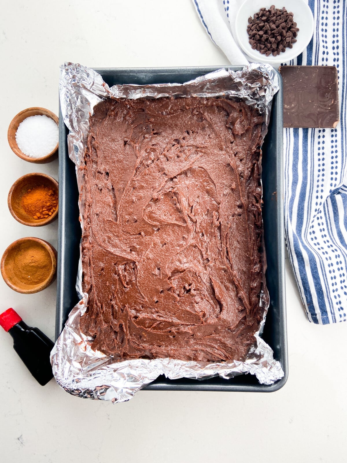 A baking pan lined with foil is filled with Mexican Brownies batter. Surrounding the pan are small bowls of salt, cinnamon, chocolate chips, a piece of chocolate, vanilla extract, and a striped kitchen towel.