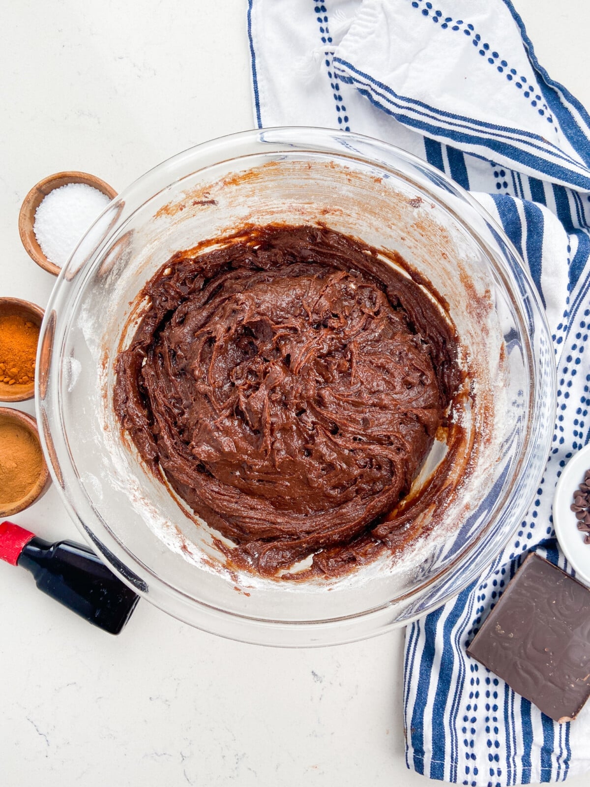 A glass bowl filled with chocolate batter for Mexican Brownies sits on a white countertop, surrounded by ingredients like salt, cinnamon, chocolate, vanilla extract, and a blue-striped kitchen towel.