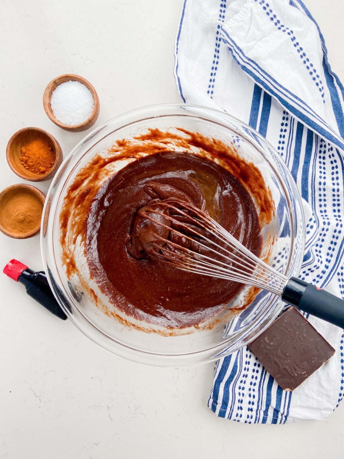 A glass bowl with chocolate batter for Mexican Brownies and a whisk sits surrounded by a blue-and-white towel, a bottle of vanilla, a bar of chocolate, and small bowls of salt, cinnamon, and another spice on a white surface.