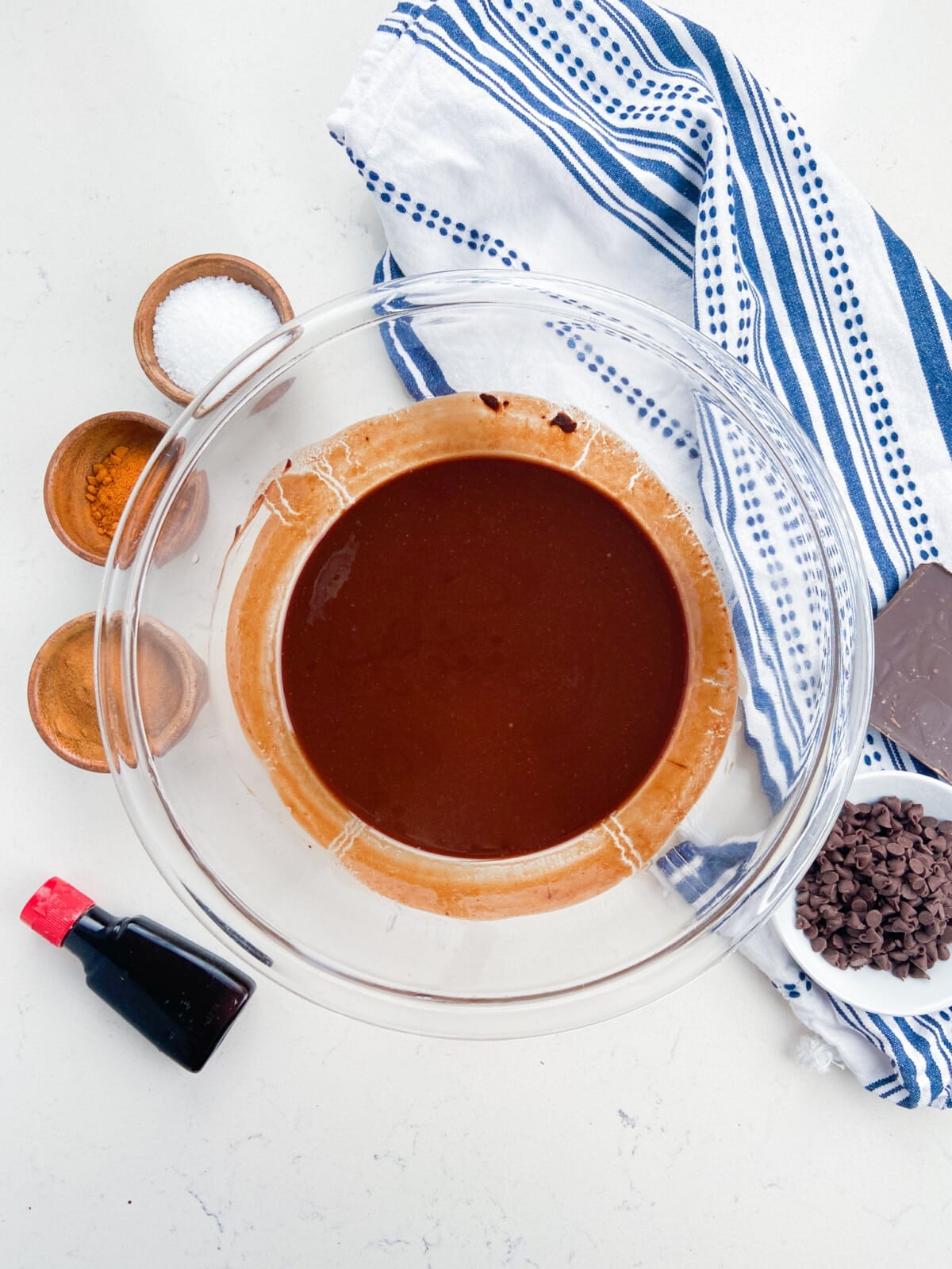 A glass bowl filled with chocolate batter for Mexican Brownies sits on a white surface, surrounded by small bowls of cocoa powder, salt, and sugar, a bottle of vanilla extract, chocolate chips, a chocolate bar, and a blue-striped towel.