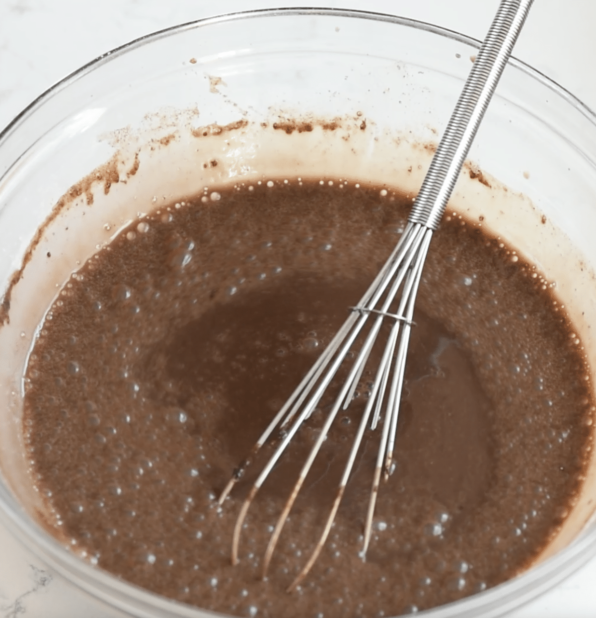 A metal whisk rests in a glass bowl filled with chocolate batter—perfect for making Slow Cooker Brownie Pudding, with bubbles on the surface and some batter splattered on the sides of the bowl.