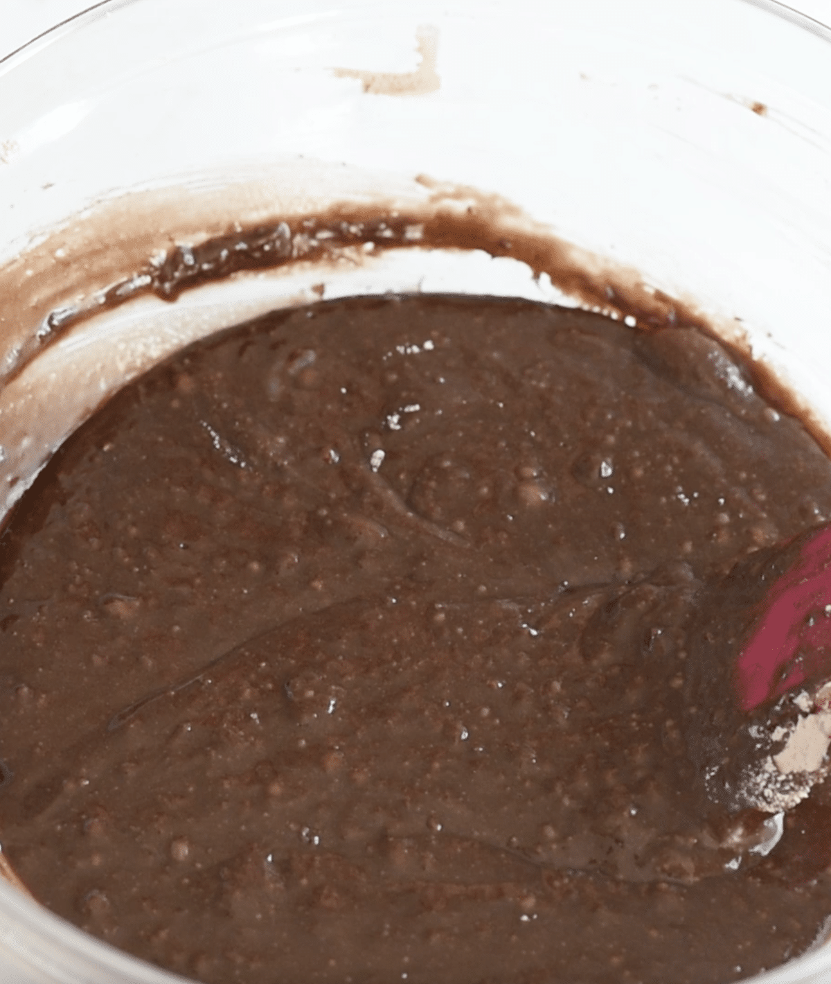 A close-up of chocolate cake batter being mixed in a clear glass bowl, with a spatula partially visible on the right. The smooth, slightly lumpy batter is perfect for making crockpot lava cake or Slow Cooker Brownie Pudding.