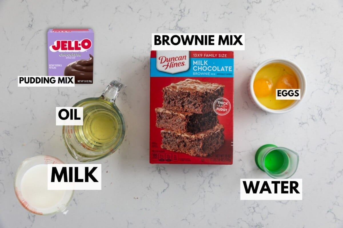 Brownie mix box, chocolate pudding mix, oil, eggs, milk, and water are arranged on a white countertop—everything you need to make crockpot lava cake or a rich Slow Cooker Brownie Pudding. Each item is clearly labeled with its name.