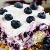 A close-up of a blueberry delight dessert slice with a crumbly crust, creamy whipped topping, blueberry filling, and whole blueberries on top.