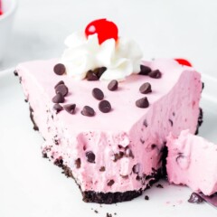 One large slice of cherry jello pie topped with chocolate chips, whipped cream and cherry with first bite missing