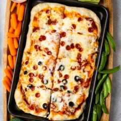 Overhead shot of sheet pan pizza on a wooden cutting board surrounded by veggies