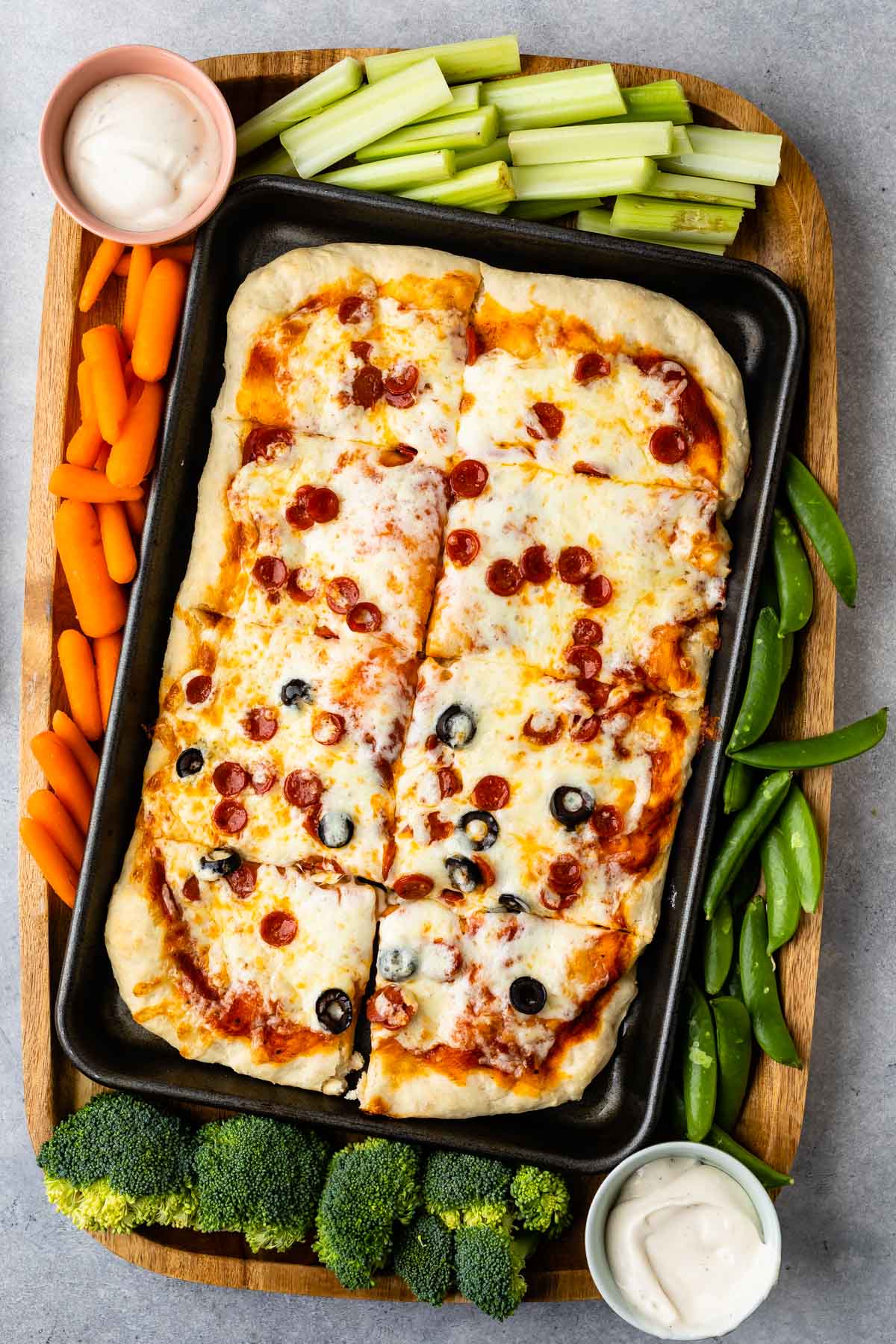 Overhead shot of sheet pan pizza on a wooden cutting board surrounded by veggies