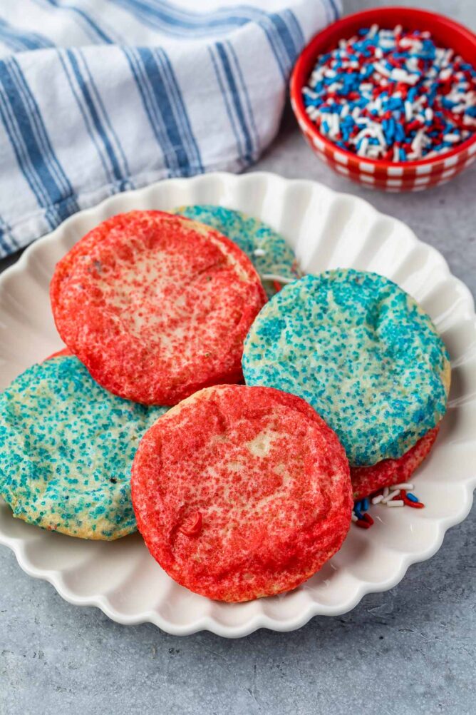 Overhead shot of blue and red sugar cookies on a plate next to a small dish of blue and red sprinkles
