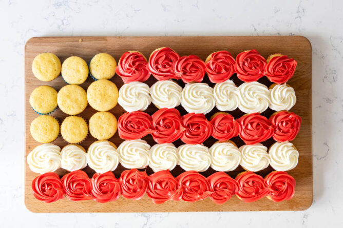 Overhead shot of vanilla cupcakes on cutting board with two rows decorated with white frosting and two decorated with red frosting