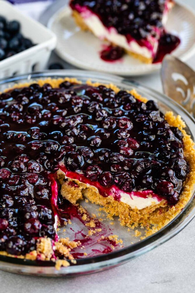 Blueberry cream cheese pie with one slice cut out and on a plate behind the pie dish
