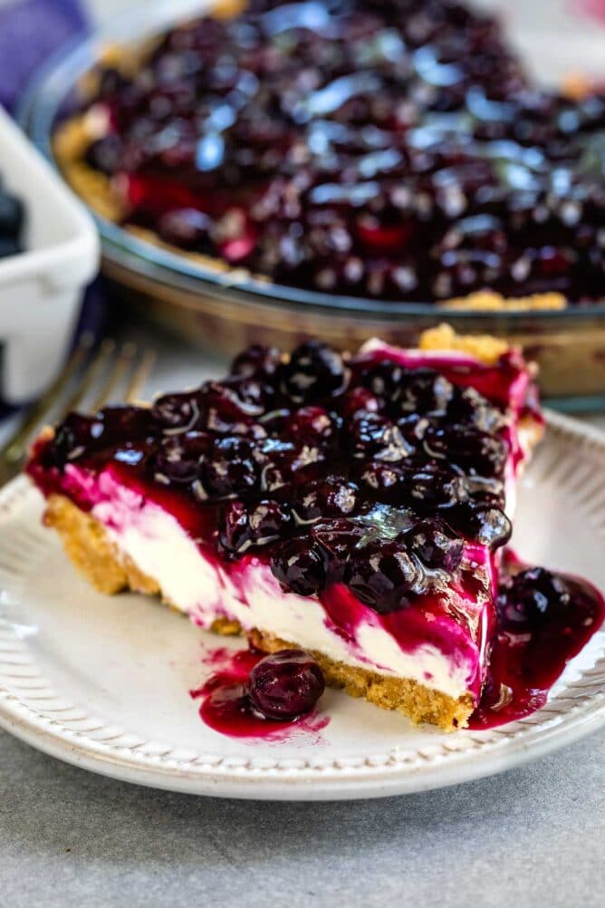 One slice of blueberry cream cheese pie on a plate with rest of pie behind it