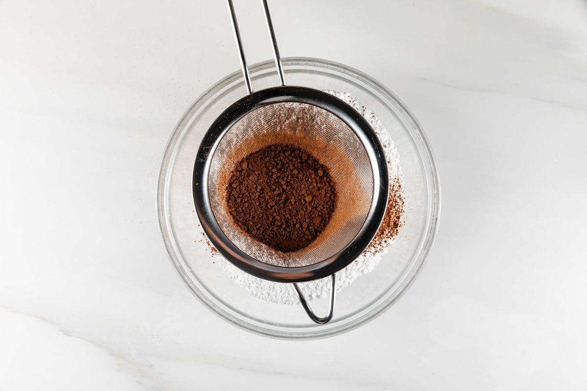 sifter sifting cocoa into powdered sugar in a glass bowl.