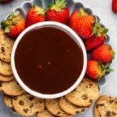 A round plate with a bowl of chocolate dip in the center, surrounded by fresh strawberries and chocolate chip cookies on a gray surface.