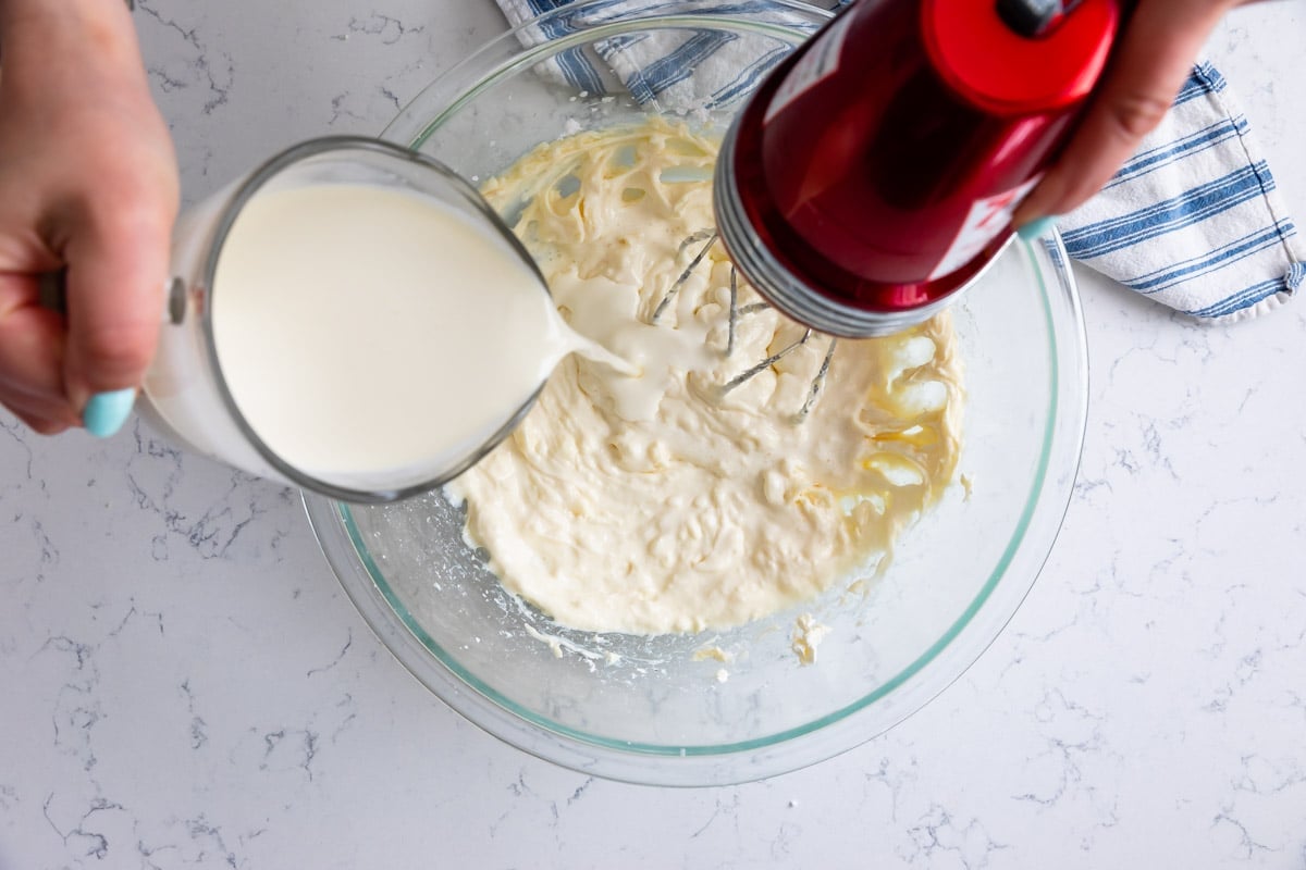 pouring cream from measuring cup into bowl with cream cheese whipe mixing with hand mixer.