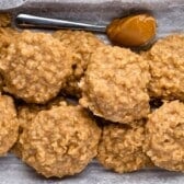 A tray lined with parchment paper holds several round, golden brown peanut butter no bake cookies with a rough, oat-filled texture. A spoon with a dollop of peanut butter rests at the top.