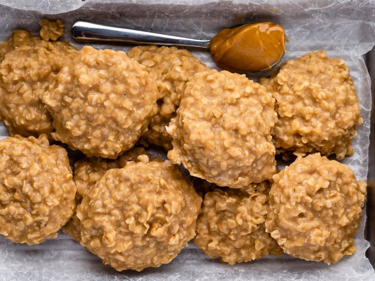 A tray lined with parchment paper holds several round, golden brown peanut butter no bake cookies with a rough, oat-filled texture. A spoon with a dollop of peanut butter rests at the top.