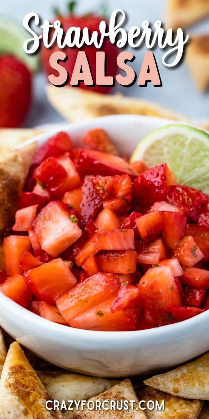 Bowl of strawberry salsa with a lime wedge surrounded by pie crust chips and recipe title on top of image