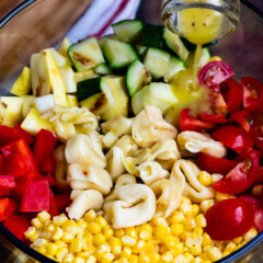 corn and zucchini and peppers and pasta and tomatoes all separated in a clear bowl with dressing being poured in