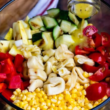 corn and zucchini and peppers and pasta and tomatoes all separated in a clear bowl with dressing being poured in