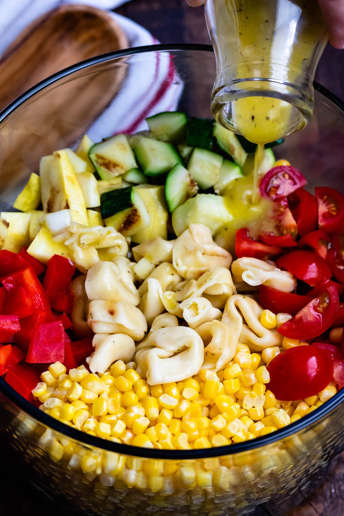 corn and zucchini and peppers and pasta and tomatoes all separated in a clear bowl with dressing being poured in