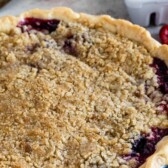 Mixed berry pie with crumble topping next to a basket of raspberries and blueberries and recipe title on top of image