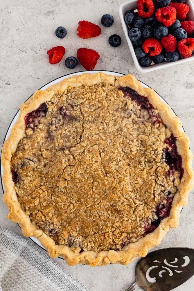 Overhead shot of mixed berry pie next to a bowl full of raspberries and blueberries