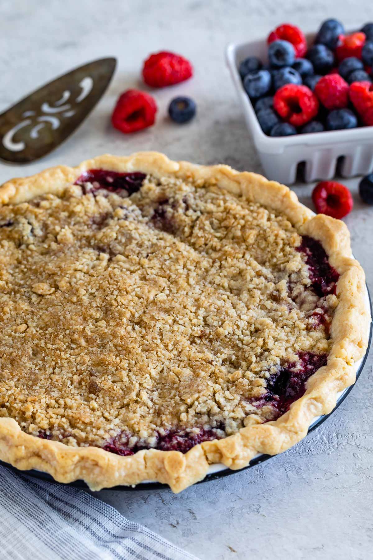 Mixed berry pie with crumble topping next to a basket of raspberries and blueberries