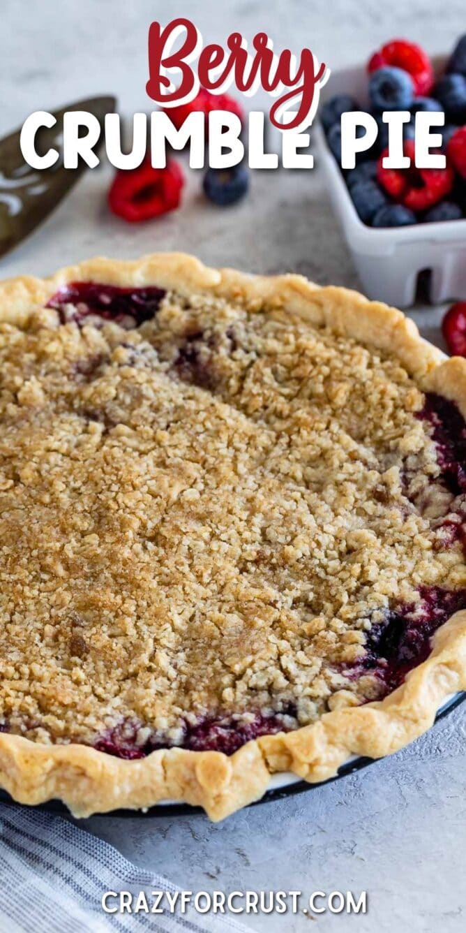 Mixed berry pie with crumble topping next to a basket of raspberries and blueberries and recipe title on top of image