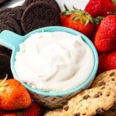 A blue bowl filled with creamy cheesecake dip is surrounded by fresh strawberries, chocolate chip cookies, and chocolate sandwich cookies on a platter.